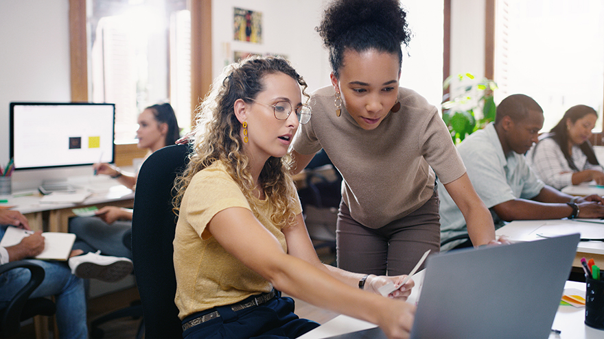 Two women collaborating at a desk with a laptop with several people working at desks in the background.